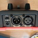 Detail zadní strany černého elektronického zařízení s červeným spodním rámem. Jsou vidět dva ovládací knoflíky, USB port a dva konektory XLR s nápisy…