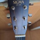 Detail hlavy akustické kytary v matně tmavě modré barvě. Na čele je zlatý nápis SIGMA, nad ním řecký znak Σ a pod ním text EST.1970. Kytara má šest c…