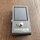 Stříbrný ladicí pedál s displejem a jedním stoupacím spínačem. Na spodní části je uveden nápis STROBOSTOMPHD™, peterson a STROBE TUNERS. Vlevo od spí…