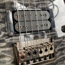 Detail elektrické kytary s černým humbuckerem a chromovaným mostem. Na snímači je viditelný nápis Seymour Duncan. Povrch kytary má šedou barvu s výra…