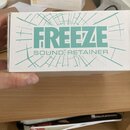 Bílá papírová krabice s tyrkysovým potiskem geometrických linií. Uprostřed je velký nápis FREEZE a pod ním menší text SOUND RETAINER. Krabice vykazuj…
