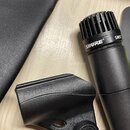 Černý dynamický mikrofon s mřížkou a nápisem SHURE® SM57. Vedle něj je černý plastový držák s vyraženým nápisem SHURE. V horní části je vidět fragmen…