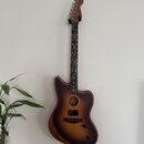 Akustická kytara s vyřezem v korpusu a tmavě hnědým přechodovým lakováním (sunburst). Na hlavě hmatníku je viditelné logo Fender. Nástroj visí na stě…