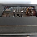 Fotografie zachycuje vnitřek černého zesilovače s otevřeným šasi. Uvnitř jsou vidět dvě elektronky, zapojení kabelů a kovové komponenty. Povrch černé…