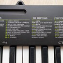 Detailní pohled na horní panel černého klávesového nástroje s bílými klávesami. Na povrchu jsou tři seznamy s nápisy 50 SONG BANK, 100 RHYTHMS a 100…