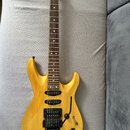 Elektrická kytara s žlutým průsvitným lakem a viditnou strukturou dřeva. Na hmatníku jsou bílé tečkové značky a na hlavě je viditelný nápis "Ibanez".…