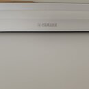 Detail horní části bílého hudebního nástroje. Uprostřed je vidět šedé logo a nápis YAMAHA. Povrch je hladký a v dobrém stavu.