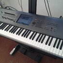 Šedý digitální klavír s černými a bílými klávesami, umístěný na černém stojanu. Na pravé straně jsou bílé nápisy KRONOS MUSIC WORKSTATION a KORG. Nás…