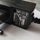 Černý síťový adaptér z plastu s připojeným kabelem a konektorem. Na štítku je uveden nápis YAMAHA, MODEL NO.: MU-18-0120150 AC ADAPTOR, INPUT: AC 100…