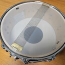 Fotografie znázorňuje snare drum z pohledu shora. Bílá membrána je napnutá na chromovaný obruč a přes ni je položen kovový síťový mechanismus s mosaz…