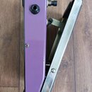 Fialový efekt pedál z pohledu z boku, vedle kterého je umístěn kovový pedalboard nebo opěrka. Na fialovém těle jsou vidět dva konektory a jeden šroub…