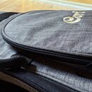 Šedý textilní obal na hudební nástroj s hrubou strukturou povrchu. V horní části je umístěno zlaté logo s nápisem Cort. Obal je vybaven černými zipy…