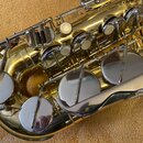 Detailní pohled na mechanismus zlatého saxofonu. Jsou vidět kovové klapy s perleťovými a kovovými krytkami. Povrch nástroje je lesklý, bez viditelnýc…