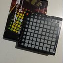Černý MIDI kontrolér Novation Launchpad Mini s mřížkou bílých podsvícených políček. Na pravém okraji je svisle napsáno LAUNCHPAD MINI a v pravém doln…