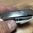 Detailní pohled na kovový, chromovaný kryt s rounded hranami, který je připevněn k bílému plastovému podkladu. Povrch chromu je výrazně poškrábaný a…