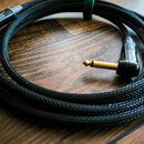 Černý audio kabel v opleteném textilním plášti, srolovaný do kruhu a sevřený zeleným stahovacím páskem. Kabel má na koncích pozlacené 6,3mm jack kone…