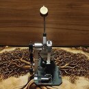 Fotografie znázorňuje kovový mechanismus pedálu, pravděpodobně pro bicí nástroj. Zařízení je stříbrné a černé s vysokou vertikální tyčí zakončenou ku…