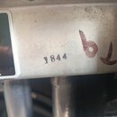 Detailní záběr na kovový povrch zařízení s viditelným opotřebením a špinavým povrchem. Na kov jsou vyražena čísla 1844 a slabší nápis F121352, vedle…