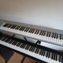 Dvě klávesové nástroje umístěné nad sebou na černých stojanech. Horní instrument je stříbrný s nápisem YAMAHA na horním panelu. Dolní instrument je č…