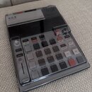 Šedý sampler s černým displejem a ovládacím panelem v průhledném plastovém krytu. Na horní části je nápis K.O.II a pod ním japonské znaky サンプラー. Pane…