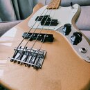 Detailní pohled na tělo elektrické basové kytary v béžové barvě s kovovým leskem. Nástroj má bílý pickguard, dva černé snímače a tři černé ovládací k…