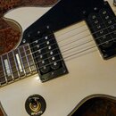 Detail elektrické kytary v krémové barvě s černým pickguardem. Nástroj má dva humbuckery s černými kryty a chromovaný kobylník. Na těle je umístěn př…