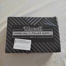 Kartonová krabice s černým povrchem a šedými šikmými pruhy. Na krabici je bílý štítek s nápisy „Movall“, „MICRO MULTI-POWER SUPPLY“ a „MPS-01“. V lev…