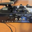 Zadní strana černého kytarového procesoru s nápisem Roland GR-55 GUITAR. Jsou vidět různé konektory s popisky GK IN, GUITAR OUT, OUTPUT R a L/MONO, P…