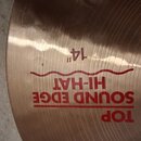 Detailní záběr na povrch mosazného činele. Na povrchu je červeným písmem nápis TOP SOUND EDGE HI-HAT a nad ním označení 14". Povrch vykazuje jemné rý…