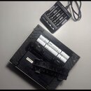 Fotografie zobrazuje černý napájecí adaptér a zadní stranu elektronického zařízení. Na adaptéru je nápis alza power. V bateriovém prostoru zařízení j…