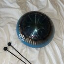 Kovový perkusní nástroj typu steel tongue drum s modrým přechodem do stříbrné barvy. Na povrchu jsou vyznačené tóny G, G, F, D, C a další symboly. K…