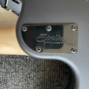 Detail zadní části šedé kytary s matným povrchem. Na těle je přišroubován lesklý kovový kryt s nápisem Sterling by MUSIC MAN. Na pravém okraji krytu…