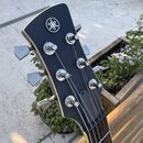Detail hlavy černé elektrické kytary s bílým lemováním. Na čele je umístěno kulaté logo Yamaha. Kytara má šest chromovaných ladiček, z nichž jedna je…