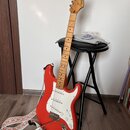 Červená elektrická kytara typu Stratocaster s bílým pickguardem a světle hnědým hmatníkem. Na hlavě kytary je nápis Squier by Fender STRATOCASTER. Ná…