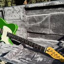 Zelená elektrická kytara s bílým pickguardem v šedém polstrovaném kufru. Na hlavě hmatníku je vidět nápis Fender Telecaster. Kytara má jeden hmatník…