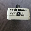 Detailní pohled na zadní stranu bílého efekt pedálu. Na šasi je černý nápis tc electronic a specifikace napájení 9 V DC 100 mA s přiloženým schématem…