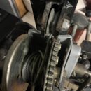 Detailní záběr na kovový mechanismus s řemenem a ozubeným kolem. Povrchy jsou šedé, vykazují známky oxidace a opotřebení. Na fotografiím nejsou žádné…