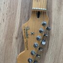 Hlava elektrické kytary z přírodního dřeva s šesti chromovanými ladičkami. Na povrchu je černý nápis Fender, STRATOCASTER a MADE IN MEXICO, dále ozna…