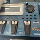 Modrý kytarový syntetizátor s nápisem GR-55 GUITAR SYNTHESIZER. Zařízení má displej, ovládací kolečko a řadu tlačítek s popisky LEAD, RHYTHM, OTHER,…