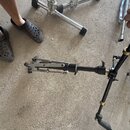Fotografie zachycuje černý kovový stojan, pravděpodobně činely nebo perkuse, položený na betonovém povrchu. Stojan má teleskopické rameno s několika…