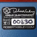 Záběr na spodní stranu modrého efekt pedálu s texturovaným povrchem. Uprostřed je černý štítek s bílým logem a nápisy KEELEY ELECTRONICS, Quality Gui…