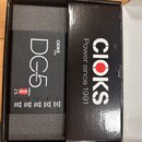Černý napájecí adaptér s nápisy DC5 POWER SUPPLY a CIOKS Link. Na spodní části jsou červené přepínače v sekci settings a nápisy 9V/12V DC 300 a 9V/12…