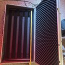 Otevřený hliníkový přepravní kufr s vnitřním polstrováním. Pravá strana víka je vyplněna černou akustickou pěnou s pyramidovým profilem, levá strana…