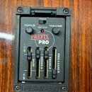 Detailní pohled na zabudovaný předzesilovač v těle akustické kytary s dřevěným povrchem. Černý ovládací panel nese nápisy NOTCH, VOLUME, PHASE a BATT…