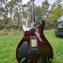 Zadní strana elektrické kytary s tmavě hnědým, lesklým povrchem s viditnou strukturou dřeva. Na zadní straně těla jsou dvě černé plastové krytky, z n…