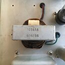 Fotografie zachycuje vnitřní součásti elektronického zařízení, konkrétně síťový transformátor s kovovým krytem. Na šedém kovovém povrchu je vyražen n…
