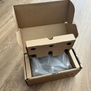 Otevřená hnědá kartonová krabice s vnitřní cardboardovou vložkou. Uvnitř je v průhledném plastovém sáčku zabalený tmavý, pravděpodobně černý, předmět…