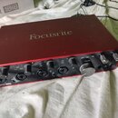 Červené audio rozhraní s nápisem Focusrite na horním krytu. Na předním panelu jsou čtyři vstupy, ovládací knofíky a velký ovratový knofík s nápisy MI…