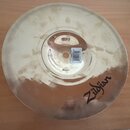Zlatavý cymbál z kovu s viditelnými šmouhama a nečistotami na povrchu. V pravé dolní části je černý nápis Zildjian. U středového otvoru je nalepená b…