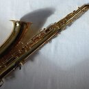 Zlatý saxofon s nablýskaným povrchem, který leží na bílém textilu. Na trubici je viditelný nápis "Selmer". Nástroj vykazuje drobné známky opotřebení…